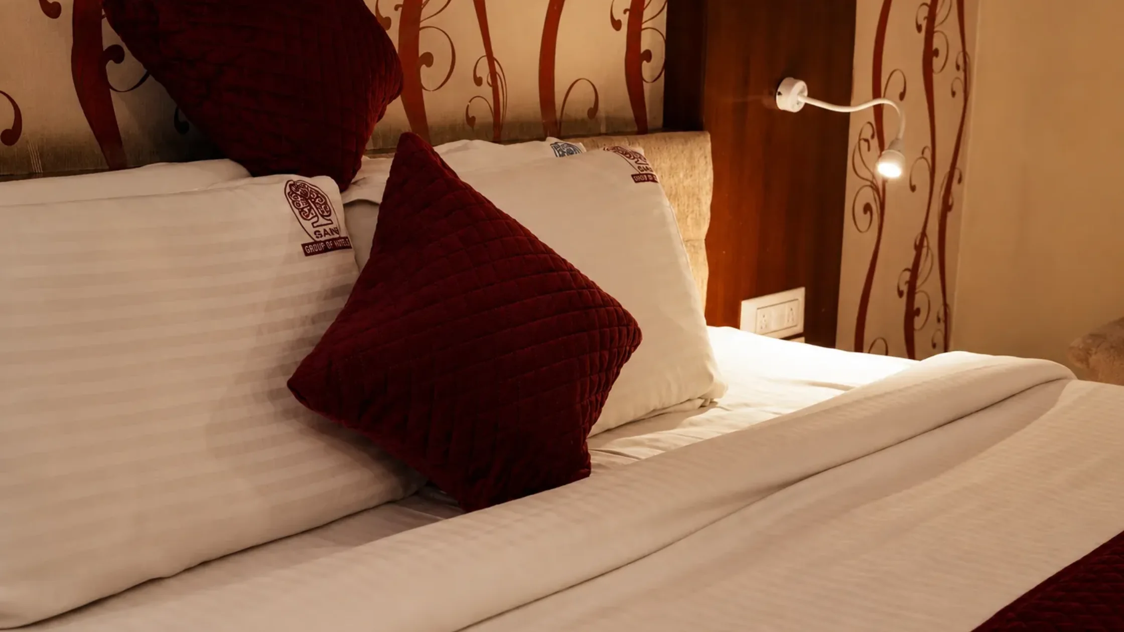 Comfortable hotel room with white bedding and maroon pillows for travellers staying near Mysore railway station and bus stand.